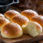 Freshly baked sourdough discard rolls, golden brown with a shiny butter-brushed top, sprinkled with flaky sea salt, displayed on a wooden board with a cozy kitchen background.