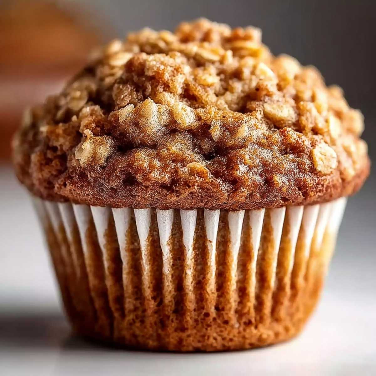 Close-up of a golden brown banana oatmeal muffin with a cinnamon crumb topping in a white paper liner, with ripe bananas blurred in the background.